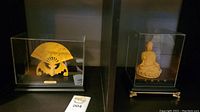 Two 24kt gold plated Asian decorative items in separate acrylic display cases on a shelf