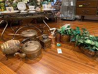 Full view of lot items arranged on wood floor beneath glass coffee table, showing kettle, barrel, chafing dish, coffee grinder, and planters with artificial plants.