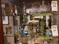 Glass cabinet shelf filled with assorted decorative items including dolls, figurines, and collectibles under tags 077 and 078.