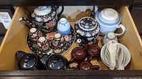 Wide view of the dishware set showing all items including floral teapots, cups, spoons, and saucers inside a wooden drawer.
