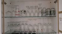 Two shelves inside cabinet with various clear glass stemware including wine, champagne, and cocktail glasses, some marked with red ink.