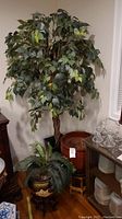 Large faux tree with broad green leaves standing behind a medium-sized faux plant in a floral painted planter on a wooden stand; large red ribbed ceramic planter nearby.