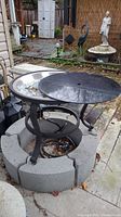 Outdoor view of the propane fireplace showing the metal round burner bowl with glass pieces inside, black metal cover, and circular concrete block base.