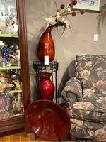 Red ceramic decorative vase set shown near a floral patterned armchair and glass display cabinet.