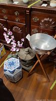 Overall view showing resin pagoda lantern, faux orchid in ceramic plant stand, and hammered metal bowl on wooden stand against wooden dresser background.