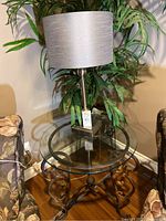 Full view of glass top table with black decorative metal base and metal lamp with gray shade on top.