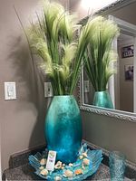 Full view of large blue iridescent glass vase with faux grass plant, placed inside blue scalloped glass bowl with sea glass and seashells.