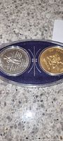 Pair of Ontario commemorative coins displayed in a transparent oval case with blue base, one silver-toned and one gold-toned, placed on a tiled surface.