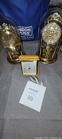 Photo showing two anniversary clocks under glass domes with gold-tone bases and a smaller rectangular clock in front, placed on grey fabric with a London Drugs bag in background