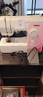 Side view of white and pink Brother sewing machine with power cord and foot pedal placed in front.