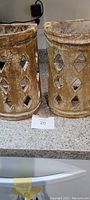 Pair of handmade ceramic cylindrical sconces with diamond-shaped cutouts, rustic tan and off-white finish, placed on a countertop.
