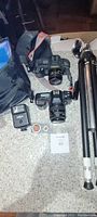 Two SLR Cameras (Yashica and Canon EOS 650) with attached lenses, Yashica branded flash, camera bag, tripod, and assorted camera accessories on table.