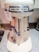 Front view of vintage 1950s Hamilton Beach malt mixer showing almond porcelain body, two mixing spindles with cross paddles, and chrome trim with Hamilton Beach nameplate.