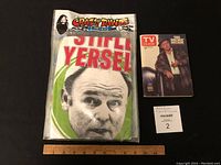 Photo of the T-shirt in original plastic bag with Archie Bunker face and text 'Stifle Yersel' visible, alongside the 1979 TV Guide with Archie Bunker on the cover and a ruler for scale.