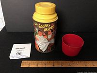 Front image of thermos showing illustration of Hogan getting a haircut, adjacent to red cup and ruler for size reference.