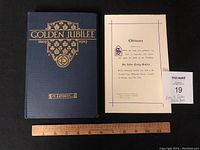 Photo of the book and obituary notice side by side with a ruler for scale, showing the book's blue cover with gold lettering and the obituary notice printed on cream paper.