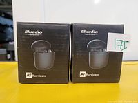 Two boxed Bluedio Hurricane wireless earbud sets in black packaging side by side on a yellow surface, displaying front view of sealed retail boxes.