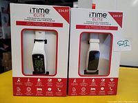 Photo of two iTime Elite Smartwatches in retail packaging, showing white silicone bands, digital displays, and features including body temperature tracker. Pricing visible on boxes.