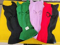 Four knit ski masks laid out side by side in black, green, purple, and red colors, showing eye and mouth cutouts and pointed ear-like tips on top.