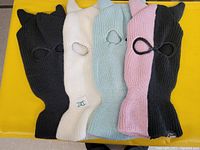 Photo shows four knit ski masks in a row on a yellow surface. Colors include black (with cat ears), white, light blue, and pink knit masks. Each mask has two eye holes and one mouth/nose hole.
