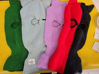 Photo shows four knit ski masks laid flat on a yellow surface. Colors are green, light gray, lavender, and a red/black two-tone mask. Each mask has a single eye hole and a mouth opening. Knit texture is clearly visible.