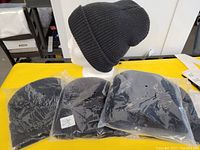 Photo of four black knit toques, individually packaged in clear plastic and displayed on a yellow surface with one on a mannequin head.
