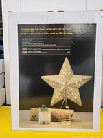 Box showing a gold glitter Christmas star tree topper with LED lights and battery pack, displayed on a black background.