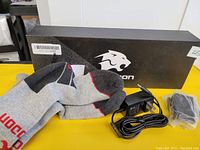 Photo showing pair of grey, black, and red heated socks laid in front of a black box with a white stylized lion logo, along with charger and batteries.