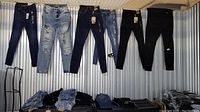 Photo showing six pairs of jeans hanging on display, including dark blue, light blue distressed, and black jeans. Below are stacks of folded jeans in similar styles and colors.