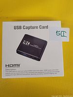 Photo of the box packaging of the USB Capture Card showing the product image and description details.