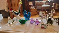 Photo of 11 various bird-shaped colorful art glass figurines on a table including a tall rooster, smaller birds in multiple colors such as blue, green, purple, and clear glass.