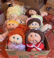 Six needle-molded Cabbage Patch Dolls placed in a cardboard box, showing faces, hair, and some clothing with visible wear and stains.