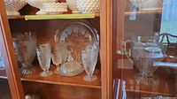 Photo showing display of the crystal iris depression glass items including goblets, covered dish, platter, and pitcher in a cabinet.