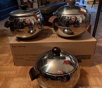 Three vintage Mid-Century Modern style penguin-themed ice buckets sitting on a cardboard box. Each is stainless steel with polished finish, wooden side handles, black knob lids, and multiple etched penguins on the surface.