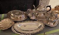 Full view of silver plated tea set including teapot, creamer, sugar, candle holders, platters, and shell-shaped bowl on black cloth near fireplace.