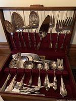 Open wooden canteen box showing silver plated serving flatware including slotted spoons, ladles, cake lifters, serving forks, and small spoons arranged in burgundy felt holder.