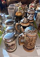 Overview photo showing eleven assorted ceramic beer steins on a table, some with lids, various decorative styles including detailed and colorful scenes.