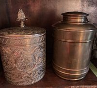Photo showing both brass canisters side by side, one etched with detailed patterns and finial lid, the other with a smooth brass finish and horizontal embossed bands.