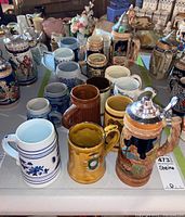 Multiple ceramic steins displayed on table showing a wide variety of styles and colors including blue and white Delft, brown Sadler steins, and some with metal lids.