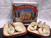 Photo showing two square cheese plates with holiday-themed spreaders and Dickens Winterville lighted house box behind.