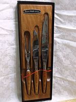 Full view of the three-piece cutlery set inserted in the wooden storage board with the utensils secured by a red cord. The utensils have polished stainless steel blades and wooden handles.