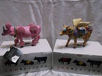 Two Westland Giftware Cow Parade figurines on white original boxes. Left is pink Angeli Cow, right is tan French Moodle cow with wings and colorful patterns.