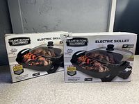 Two new Granitestone Diamond 12-inch electric skillets in original boxes, showing front packaging with image of skillet and lid containing cooked food, positioned on countertop against wall.