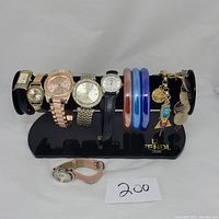 Jewelry lot showing 6 watches on a black Fendi bracelet display stand with 3 glass bangles and 2 charm bracelets on the right, plus one loose watch with pink fabric strap in front.