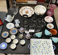 Overview of all assorted china and glassware items arranged on a black table including teapots, cups, saucers, glass bowls, pitchers, and a serving tray.