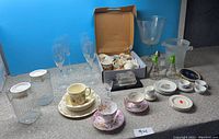 Full view of various china and glassware items arranged on floor against blue backdrop including cups, plates, tall glasses, decanters, ice bucket, and condiment set.