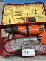 Orange Hilti electric percussion drill in a red metal box, surrounded by yellow informational charts about drill bits and anchors.