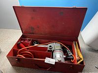 Red metal carrying case open showing Hilti Torna 765 hammer drill, orange power cord, red auxiliary handle, and accessories inside compartments.