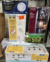 Photo showing boxes of four lighting fixtures: Hampton Bay motion sensing exterior wall lantern, Home Decorators Collection large exterior wall lantern, Hampton Bay 4-light directional track fixture, and LED landscape light kit.