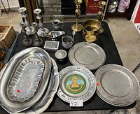 Photo showing various pewter trays, plates, a colorful USA emblem plate, candle holders, and brass decor on a table.